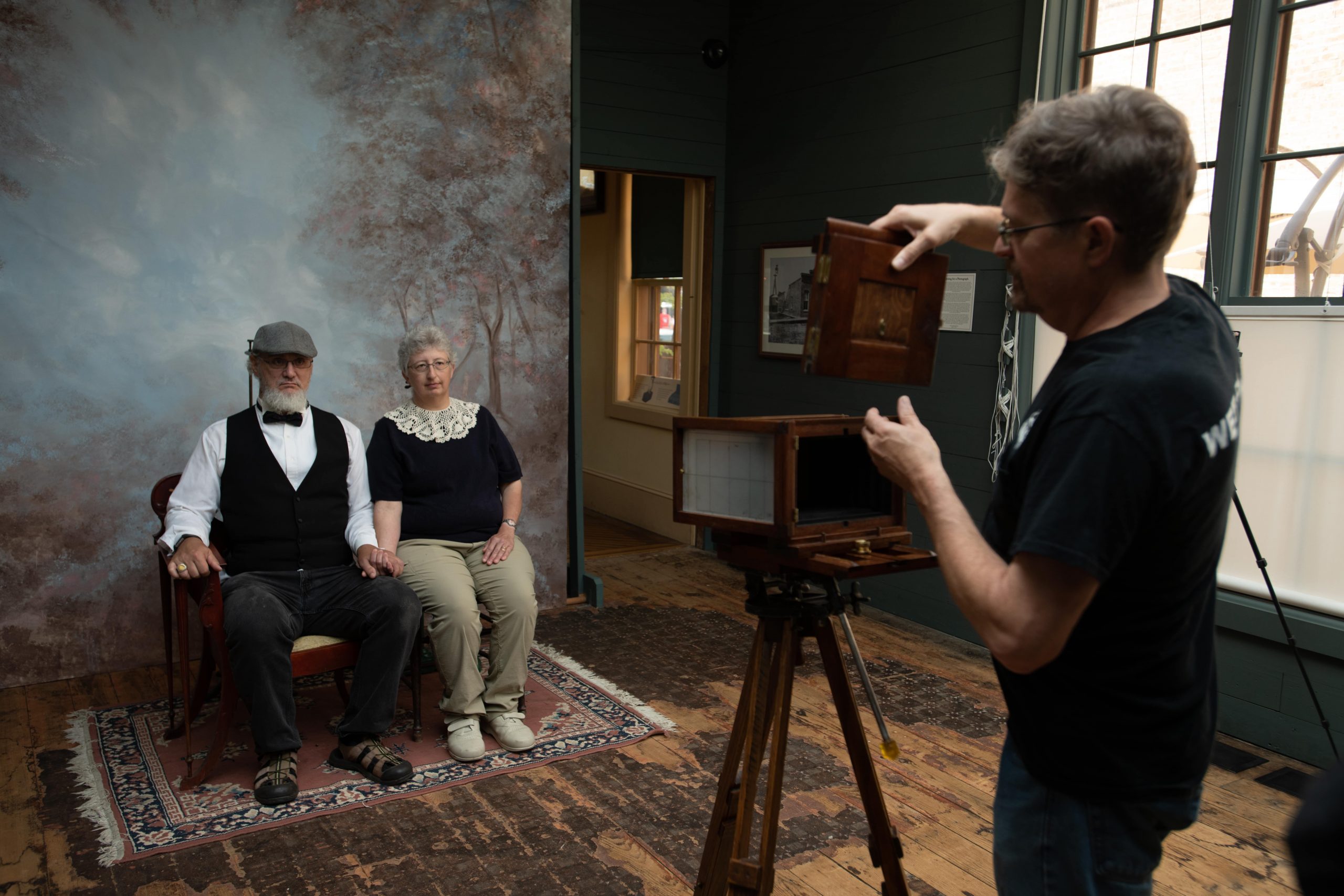 Dave prepares to take photo to create  tintypes of an older couple sitting and holding hands in mildly old fashioned clothing. 