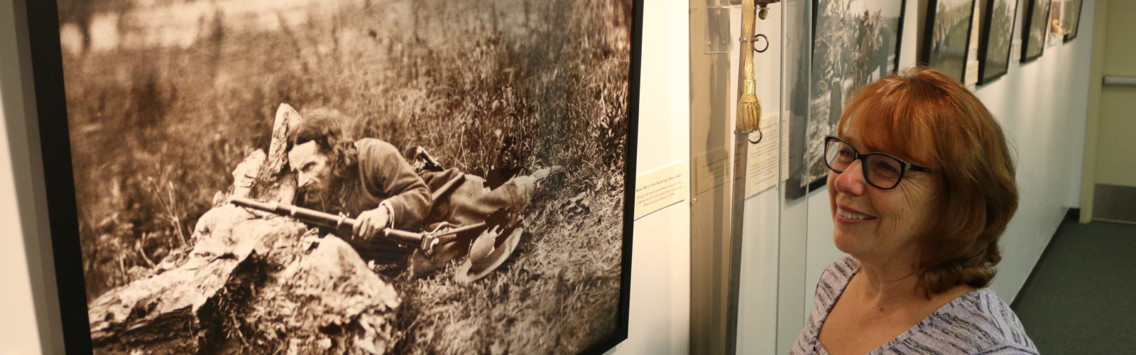 a smiling woman looks at a photo in a gallery of a soldier hiding behind a rock