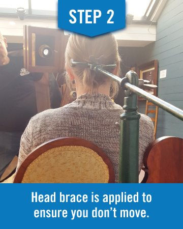 A woman sits with her head braced, viewed from behind the image reads "step 2 head brace is applied to ensure you don't move"