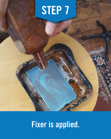 Someone pours a liquid substance over the negative appearing image in a metal tray, this image reads "step 7 fixer is applied"