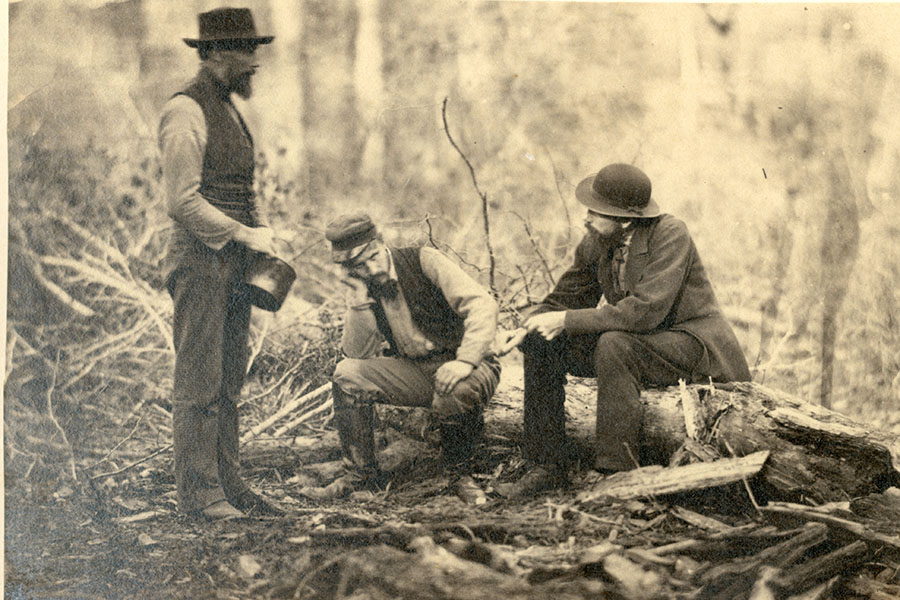 a black and white photo of two men sitting on a log while a thirds stands in front of them