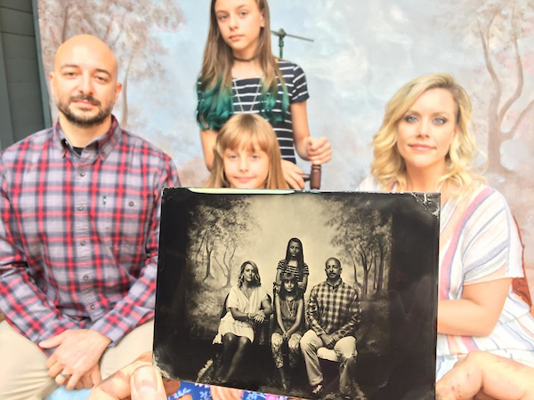 A 4 person family (husband, wife, 2 daughters) pose for their tintypes, featured in the foreground.